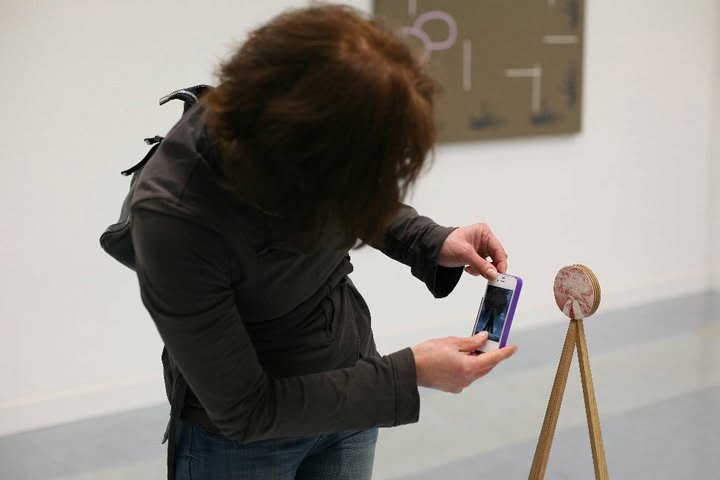 A visitor photographs a small circular artwork mounted on a wooden tripod stand