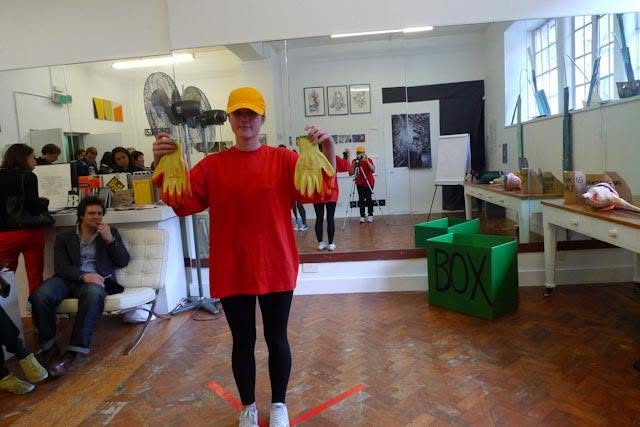 Sluice Art Fair — a performer in a red shirt and yellow cap holding yellow rubber gloves before an audience, beside green crates marked BOX