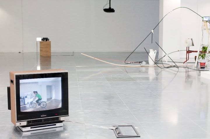 A television playing a film of a person cycling, with wire and rod sculptures behind