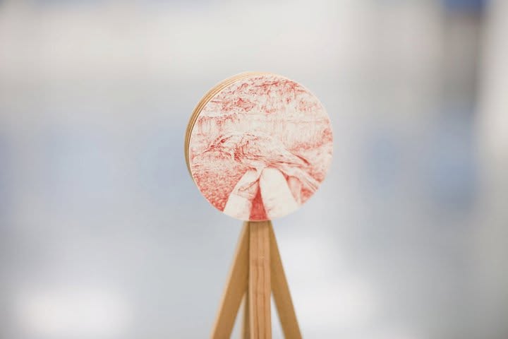 A circular wooden plaque with a red drawing of a bird held in fingers, on a tripod stand