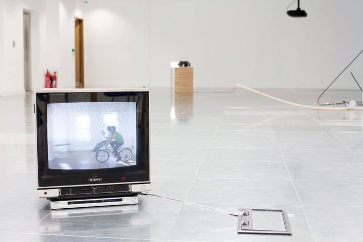 A television on the gallery floor playing a film of two people cycling