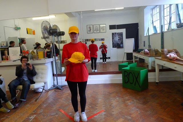 Sluice Art Fair — the performer holding a yellow cap before the seated audience, beside a green cube marked BOX